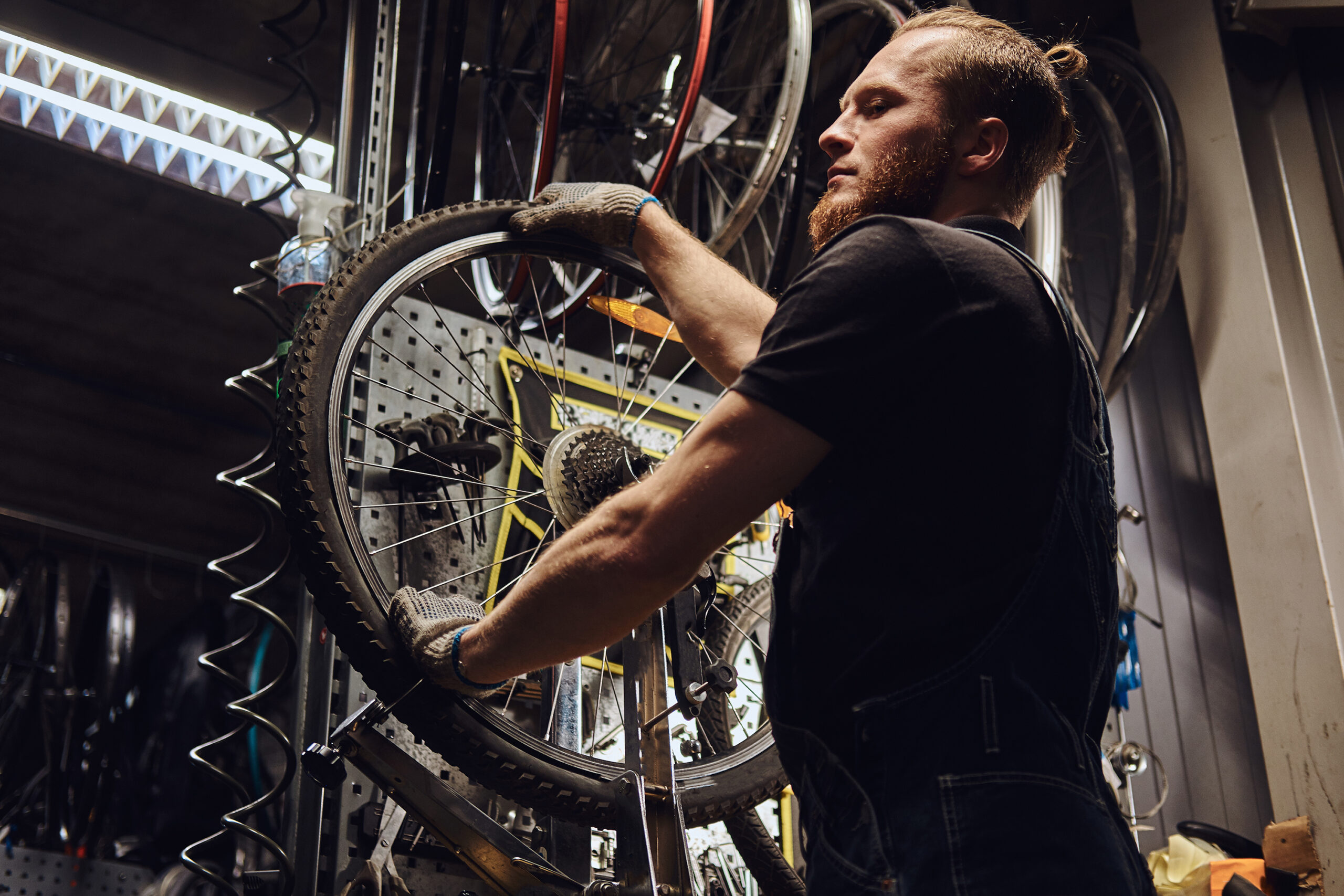 A handsome redhead male in a jeans coverall, working with a bicycle wheel in a repair shop. A worker removes the bicycle tire in a workshop.